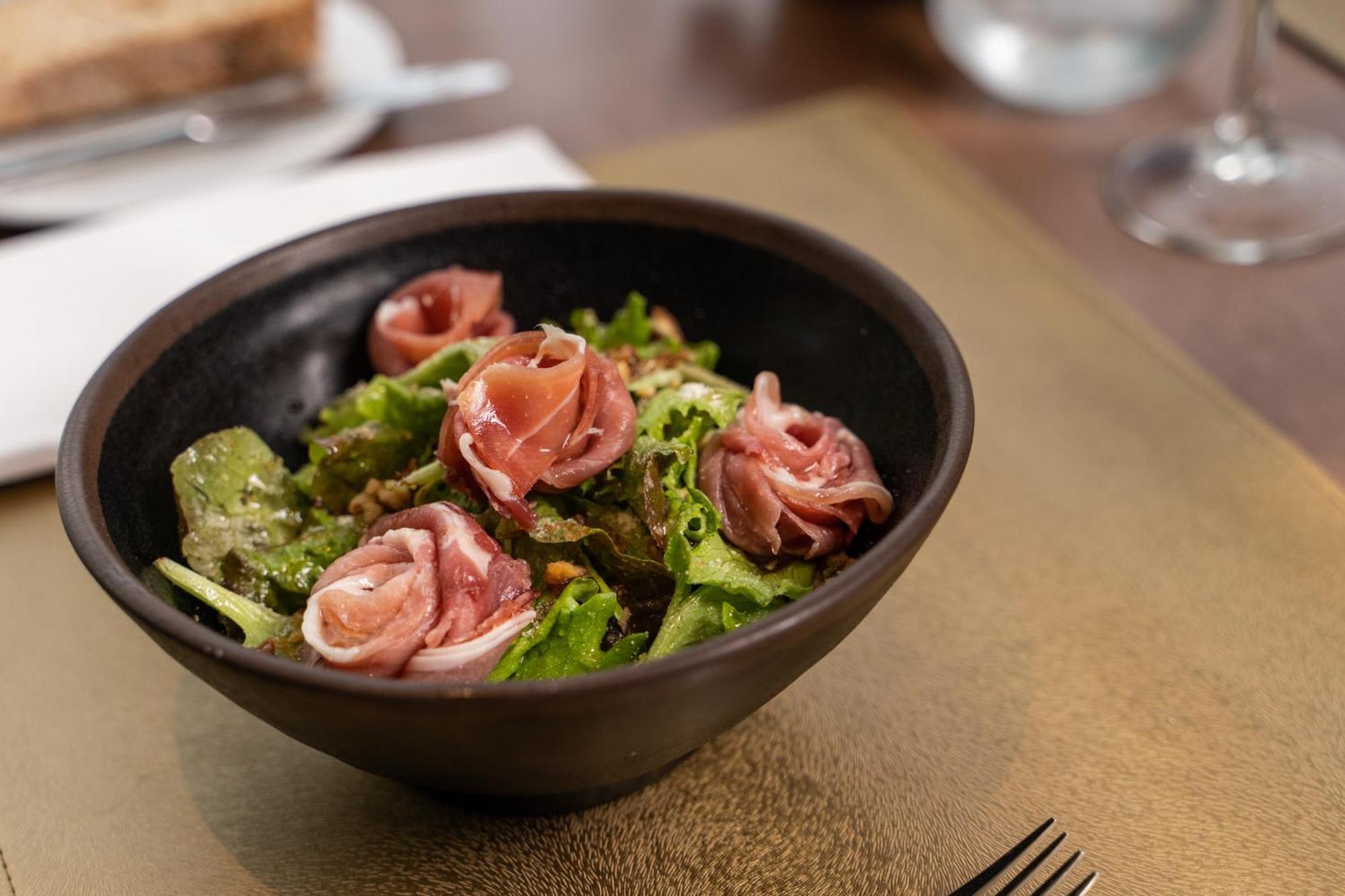 Eurostars Universal Lisboa מראה חיצוני תמונה The photo shows a bowl of salad with a mix of fresh greens, topped with rolled slices of cured meat, likely ham. The salad has a vibrant color, featuring leafy greens and the pinkish hue of the meat. The bowl is dark and round, with a simple, elegant