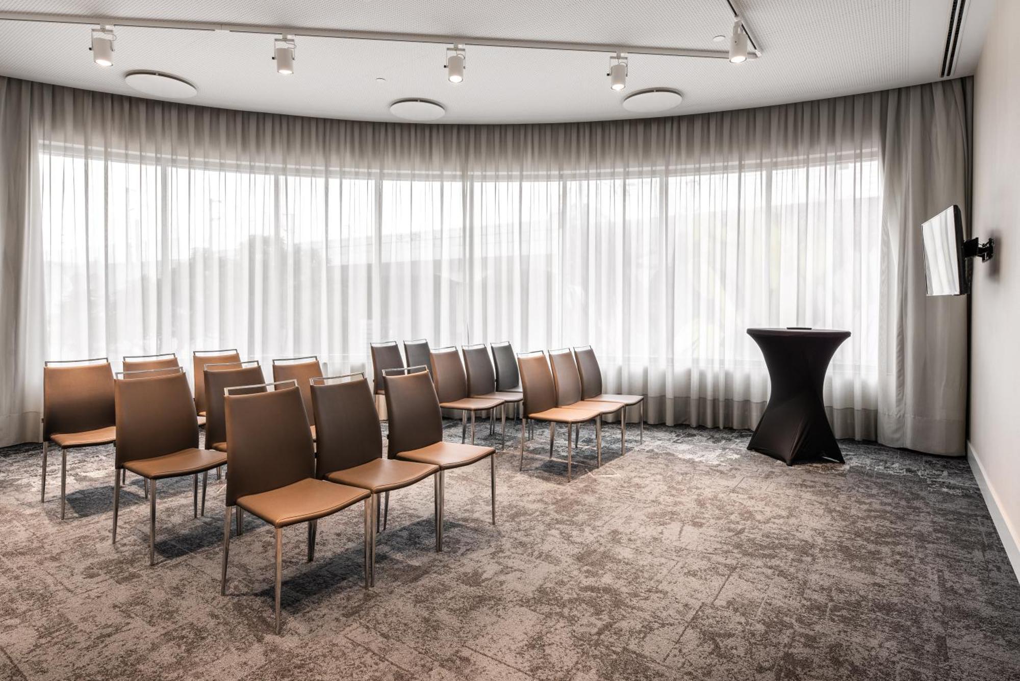 Eurostars Universal Lisboa מראה חיצוני תמונה The photo shows a modern meeting room with a curved design. There are several rows of simple, brown chairs arranged in the space, facing a small black podium. The room features large windows covered with sheer curtains, allowing natural light to filt